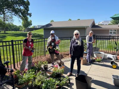 Four women stand together outdoors on a sunny day at the Jesse Helms Nursing Center, while holding plants and gardening supplies. Trees, a black fence, and patio seating are visible in the background during a community gardening project.