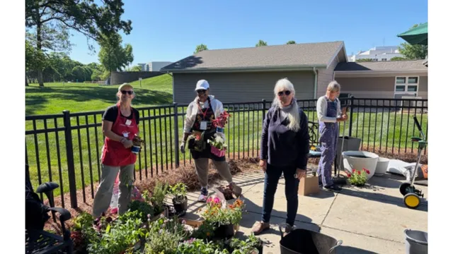 Four women stand together outdoors on a sunny day at the Jesse Helms Nursing Center, while holding plants and gardening supplies. Trees, a black fence, and patio seating are visible in the background during a community gardening project.