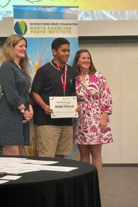 teen youth posing with 2 women in front of NC Youth Institute banner, holding a "Rising Scholar" certificate
