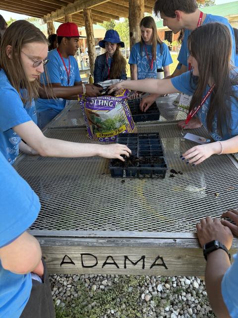 Many youth students in blue tshirts standing around a table with a metal grate, seeding soils and seed trays