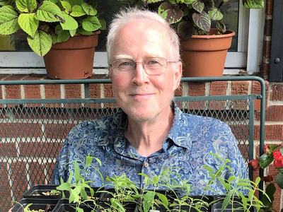 John Bowen, a smiling older man, is pictured surrounded by healthy plants he has propagated.