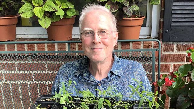 John Bowen, a smiling older man, is pictured surrounded by healthy plants he has propagated.