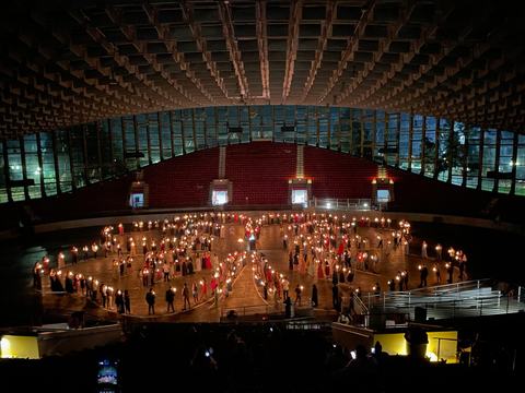 Overhead view of dozens of people holding lit candles, arranged in the shape of the 4-H emblem on the floor of a large indoor arena beneath a vaulted ceiling, audience seating in shadow.
