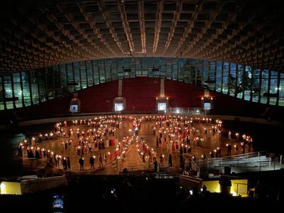 Overhead view of dozens of people holding lit candles, arranged in the shape of the 4-H emblem on the floor of a large indoor arena beneath a vaulted ceiling, audience seating in shadow.