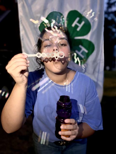 A person in a blue shirt blows soap bubbles through a wand while holding a purple bubble bottle, standing in front of a 4-H clover banner.