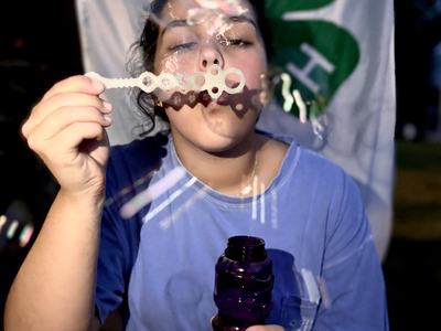A person in a blue shirt blows soap bubbles through a wand while holding a purple bubble bottle, standing in front of a 4-H clover banner.