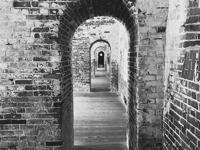Black-and-white photo of a long brick corridor with repeating arched doorways receding into the distance, wooden floorboards and textured masonry walls creating a strong sense of depth and rhythm.