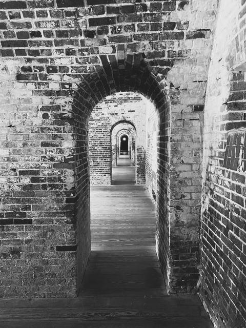 Black-and-white photo of a long brick corridor with repeating arched doorways receding into the distance, wooden floorboards and textured masonry walls creating a strong sense of depth and rhythm.