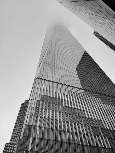 Black-and-white low-angle view of a glass skyscraper disappearing into fog, its reflective facade and vertical ribs stretching skyward; neighboring towers and geometric patterns emphasize height and scale.