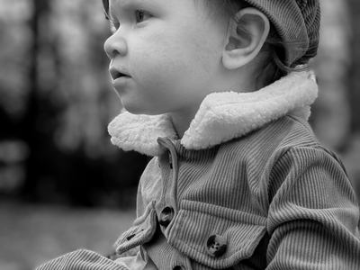 Black-and-white portrait of a toddler outdoors, wearing a corduroy coat with a shearling collar and matching beret, gazing thoughtfully to the left against blurred trees.