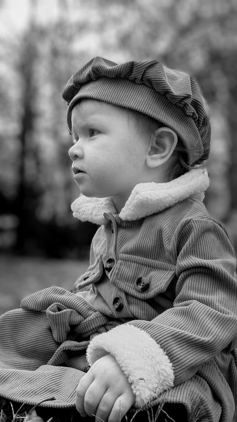 Black-and-white portrait of a toddler outdoors, wearing a corduroy coat with a shearling collar and matching beret, gazing thoughtfully to the left against blurred trees.