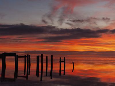 Silhouetted wooden pilings of a broken pier stand against a fiery orange and purple sunset, their dark reflections mirrored in the calm water beneath dramatic streaked clouds.