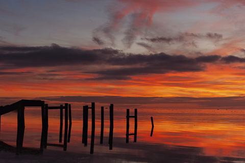 Silhouetted wooden pilings of a broken pier stand against a fiery orange and purple sunset, their dark reflections mirrored in the calm water beneath dramatic streaked clouds.