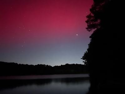 Quiet lake at night with a dark treeline silhouette; a vivid red-pink sky glow and scattered stars rise above the horizon, a bright point reflecting faintly on the water.