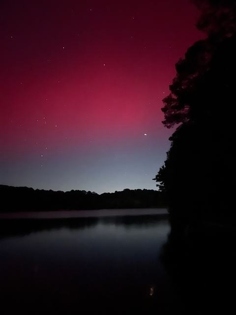 Quiet lake at night with a dark treeline silhouette; a vivid red-pink sky glow and scattered stars rise above the horizon, a bright point reflecting faintly on the water.