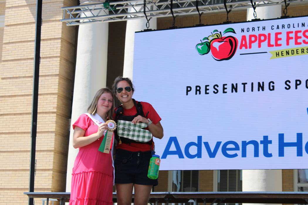Two women stand side buy side on the Apple Festival Stage. One is wearing a pink dress and holding a prize ribbon. One is holding a gingham oven mitt. They are both smiling.