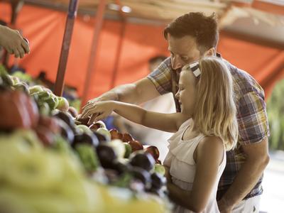 A father and young daughter examine fresh produce at a bright and sunny farmers market stall.