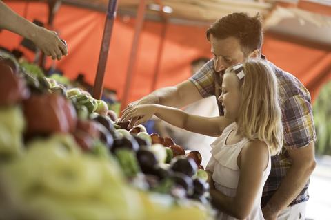 A father and young daughter examine fresh produce at a bright and sunny farmers market stall.