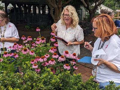 Three women holding clipboards look at purple and pink flowers