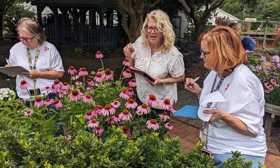 Three women holding clipboards look at purple and pink flowers