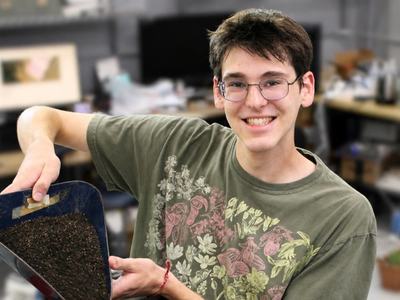 NC State student Michael Schruckmayr smiles while holding a scoop of soil and seeds in a research lab.