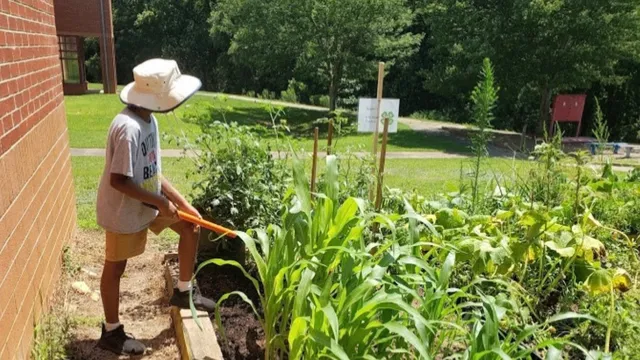 A young person wearing a sun hat tends to a lush raised-bed garden next to a brick building.