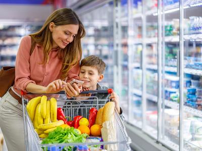 A mother and her young son use a phone to calculate the cost of food items while standing behind a grocery cart with fresh produce and bread inside.