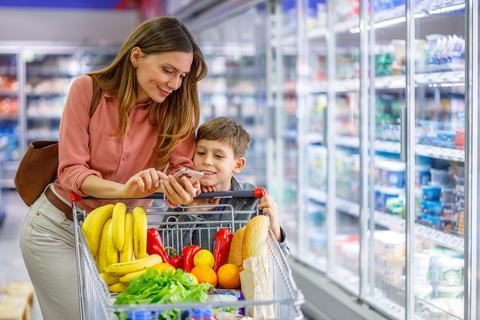 A mother and her young son use a phone to calculate the cost of food items while standing behind a grocery cart with fresh produce and bread inside.