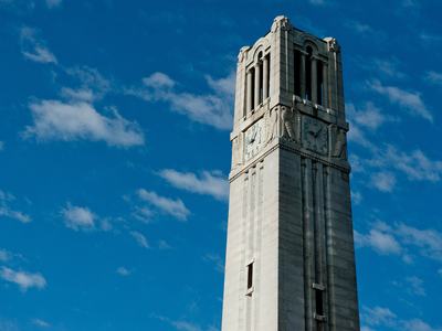 The NC State Belltower clock on a sunny day with a bright blue sky behind it.