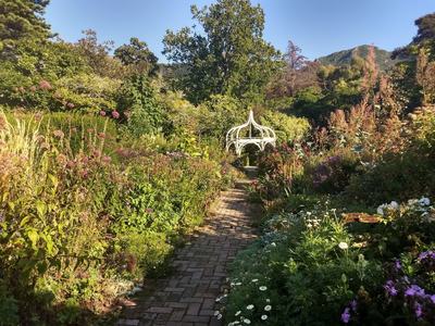 A lush garden with a white pergola at its center is surrounded by rugged hills.