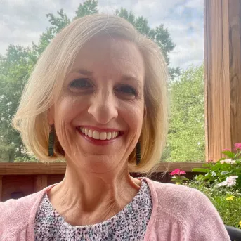 Woman sitting on a screened porch smiling with flowers and trees in the background