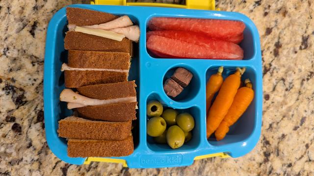 bento style lunch container with a sandwich, watermelon, carrots, and olives.