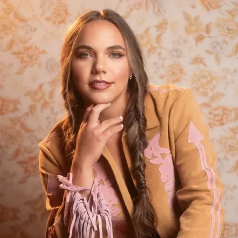 Portrait of Paige King Johnson wearing a tan jacket with pink western-style accents, resting her chin on her hand and smiling against a warm floral background.