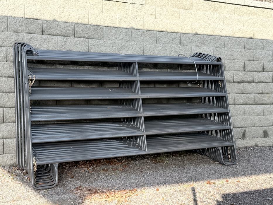 A stack of several heavy-duty, dark gray metal corral panels leans against a light gray textured stone wall.