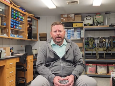 Man in a gray hoodie sits in a well-organized workshop, surrounded by tools and equipment on shelves. The setting appears practical and focused.