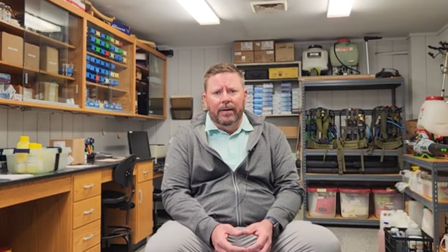 Man in a gray hoodie sits in a well-organized workshop, surrounded by tools and equipment on shelves. The setting appears practical and focused.