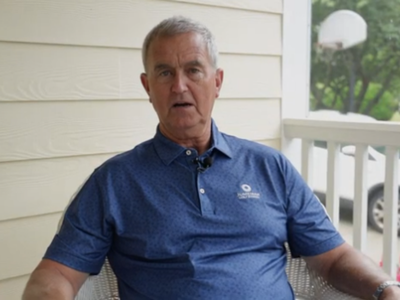 An older man sits on a porch wearing a blue polo shirt, looking into the camera with a neutral expression. A basketball hoop and greenery are visible outside.