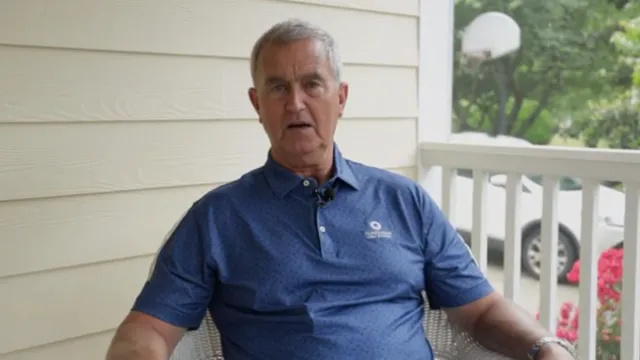 An older man sits on a porch wearing a blue polo shirt, looking into the camera with a neutral expression. A basketball hoop and greenery are visible outside.