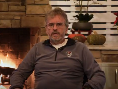 Man in a grey pullover sits in front of a lit fireplace. He appears relaxed. Background includes festive decor: red poinsettia and a pumpkin. Cozy ambiance.