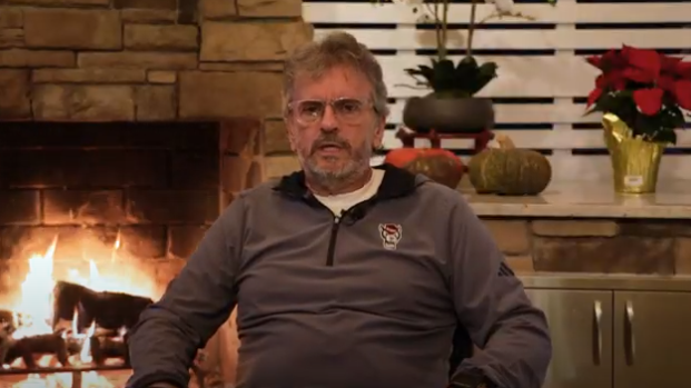Man in a grey pullover sits in front of a lit fireplace. He appears relaxed. Background includes festive decor: red poinsettia and a pumpkin. Cozy ambiance.