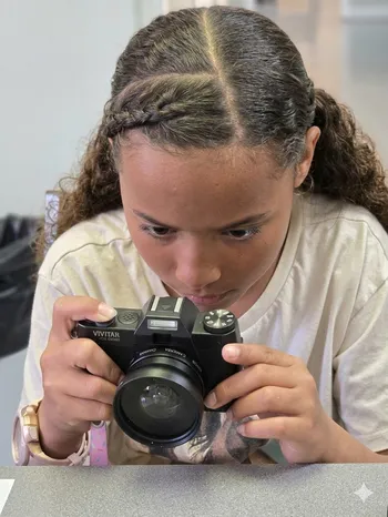 A close-up photograph focuses on a young person with curly brown hair parted and intricately braided on top. She is leaning over a table and looking intensely through the viewfinder of a black digital camera held with both hands.
