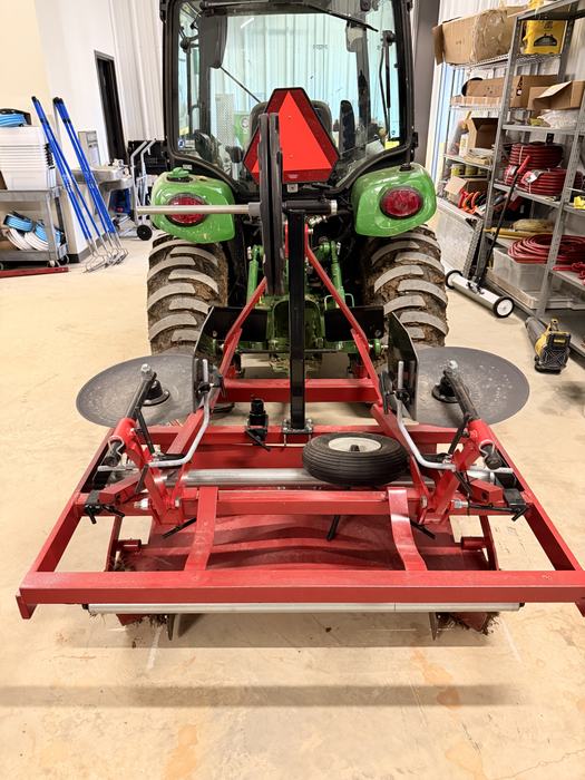 A red plastic mulch layer attachment is hitched to the back of a green John Deere tractor inside a workshop.