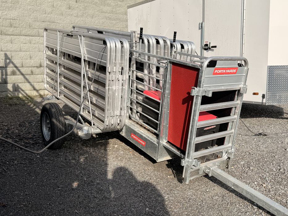 A compact, silver aluminum Porta Yards mobile livestock pen system is shown folded for transport on a trailer. The unit features red accents and a stack of interlocking gates, parked on a gravel surface next to a white trailer and a brick wall.