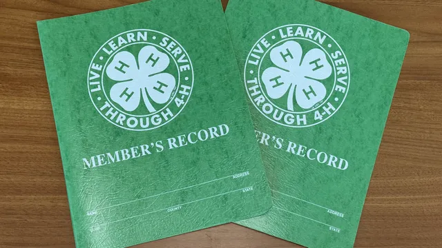 Two green 4-H project record books with white 4-H emblems and the motto "Live • Learn • Serve • Through 4-H" overlap on a wooden table.