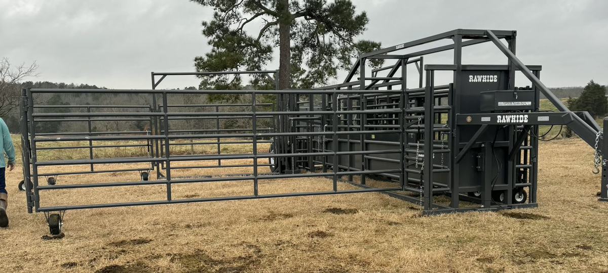 A large, dark gray "Rawhide" portable cattle corral system is set up in a dry, grassy field under a cloudy sky. The metal structure features various gates, panels on wheels, and a loading chute. A person's legs are visible on the far left.