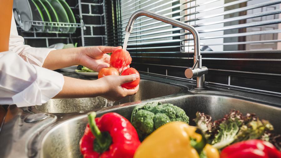 A woman rinses tomatoes under running water in a kitchen sink, while a basket of other fresh fruits and vegetables awaiting rinsing, like broccoli and bell peppers, is seen to the side.