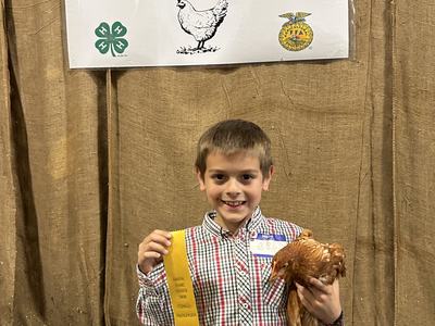 Samson Mills holding his chicken and participation ribbon in front of the Coastal Plains Chicken Show banner