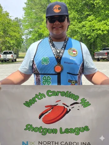 A young person with dark hair, sunglasses, and a baseball cap. He is standing in a gravel parking area with trees and vehicles in the background, holding up a large white banner with the words "North Carolina 4-H Shotgun League."