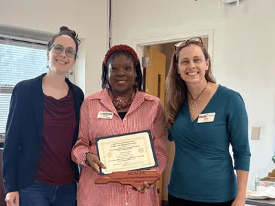 Three women posing next to each other in a classroom with the women in the middle holding award.