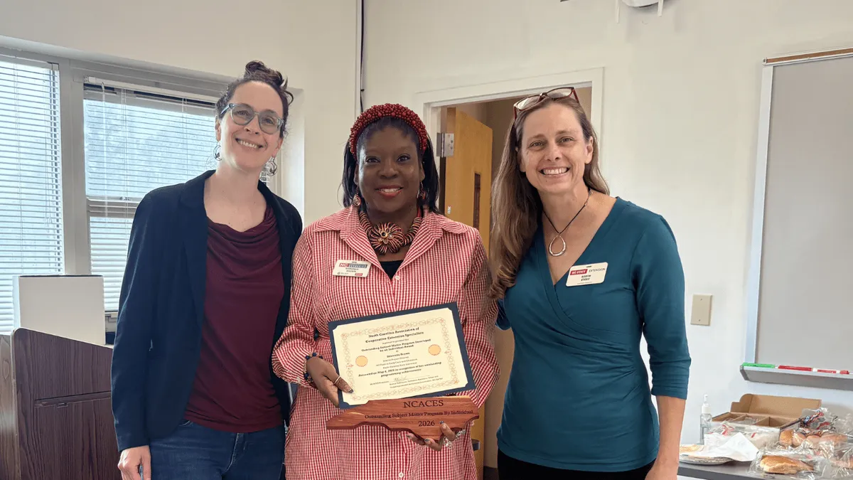 Three women posing next to each other in a classroom with the women in the middle holding award.